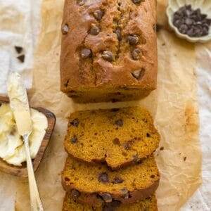 A loaf of chocolate chip pumpkin bread with three slices and a bowl of chocolate chips and butter beside it.