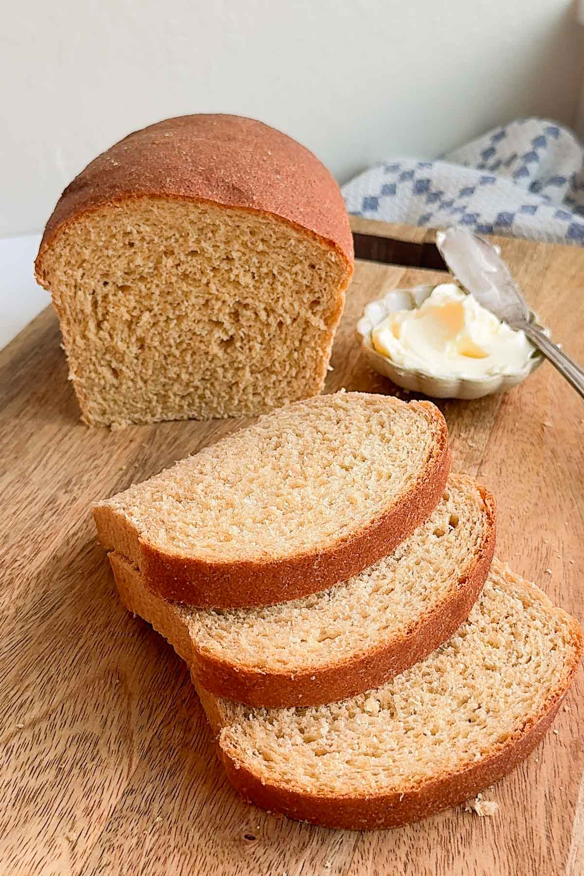 A loaf of honey whole wheat bread on a cutting board, with a small bowl of butter and three slices of bread.