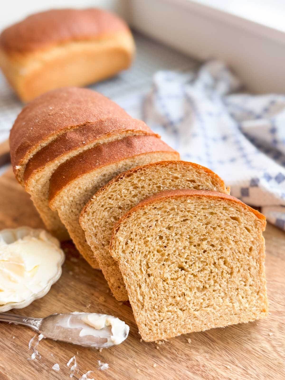 Sliced honey whole wheat sandwich bread on a wood cutting board with a small bowl of butter.